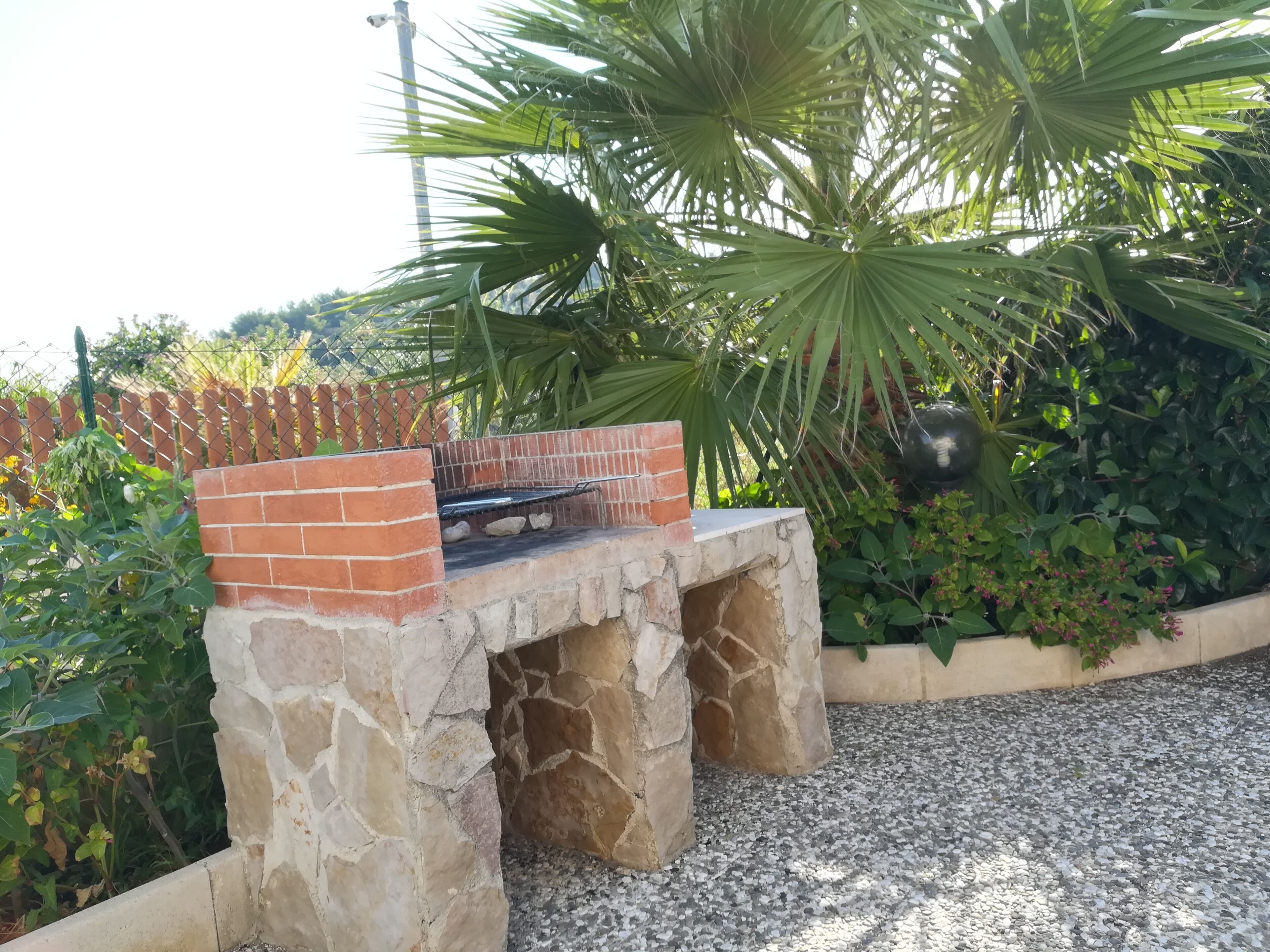 A stone-built outdoor grill with a brick chimney, surrounded by greenery and a gravel path.
