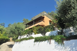 A two-story building with a sloped roof, surrounded by lush greenery and a white wall.