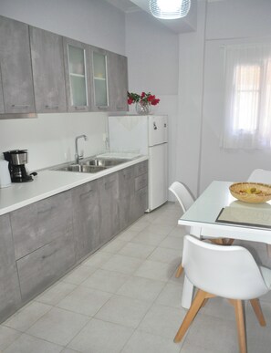 A modern kitchen with grey cabinets, a white countertop, a sink, a coffee maker, and a refrigerator. There is a dining table with chairs and a basket of fruit.
