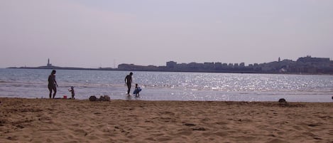 A beach with sand and water, a distant city skyline, and a lighthouse.