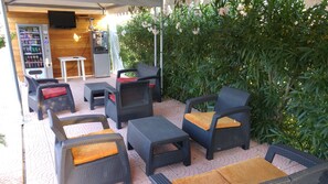 Ett utomhusområde med sittplatser och möbler i rotting, en automattank för drycker och en tv-skärm som är monterad på väggen.