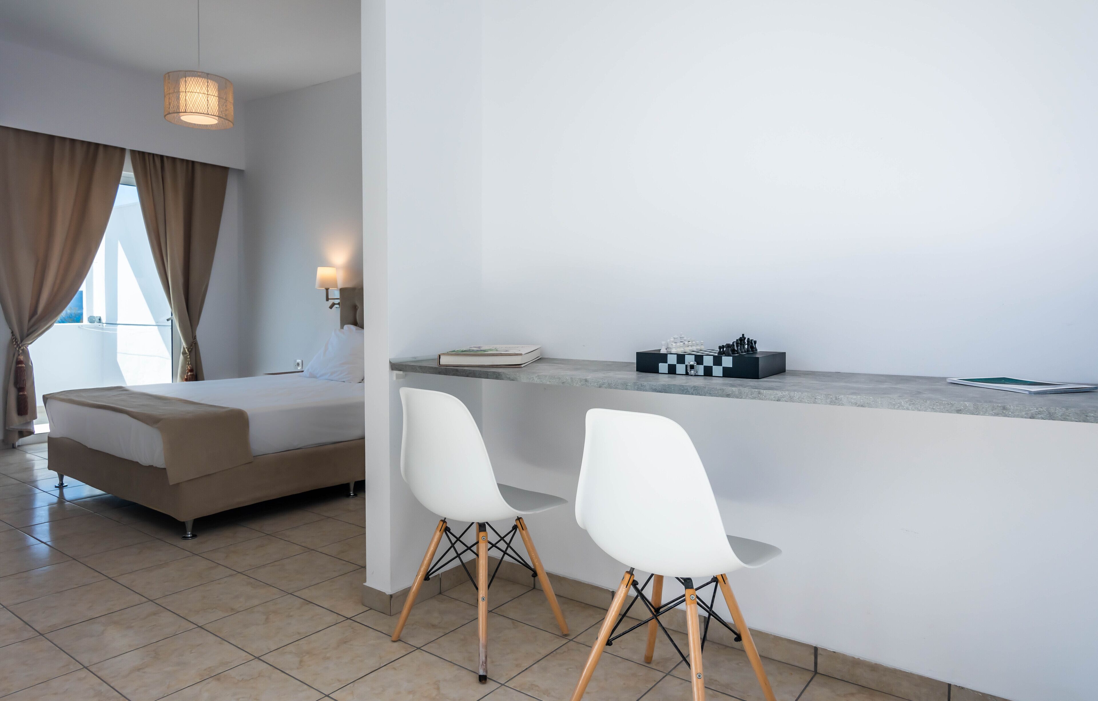 A modern hotel room with a bed, two white chairs, a small table, and a chessboard.