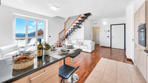 Eine moderne Küche mit einer zentralen Kochinsel, ein Essbereich mit weißem Tisch und Stühlen, eine Treppe und ein Blick ins Freie.