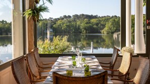 A dining table set for four with wicker chairs, a view of a lake, and a plant on the window sill. - Brindos Lac & Château (Anglet)