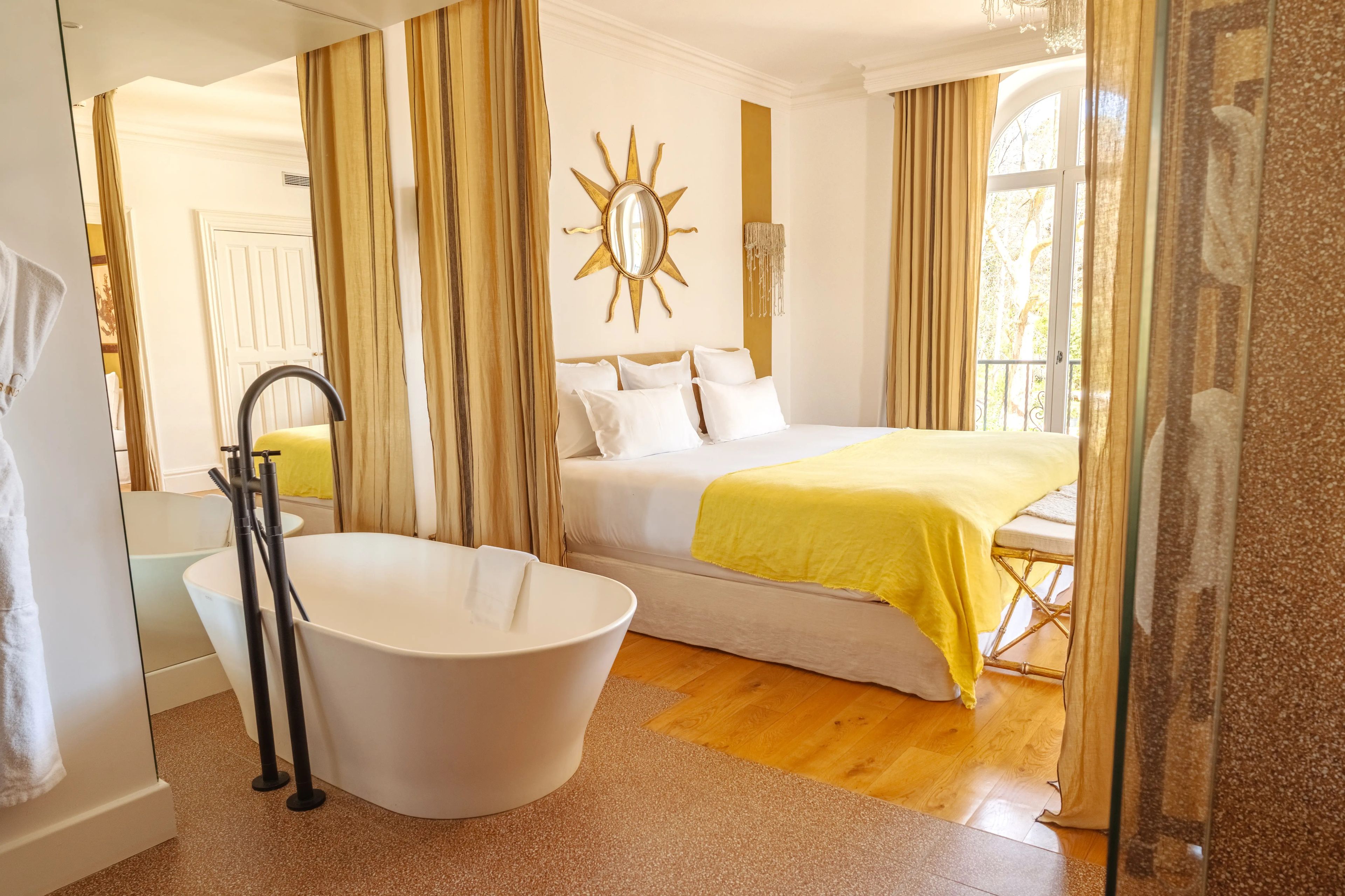 A hotel room with a bathtub, a bed with a yellow blanket, a sun-shaped mirror, and a window with curtains.