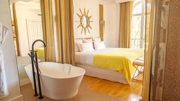 A hotel room with a bathtub, a bed with a yellow blanket, a sun-shaped mirror, and a window with curtains.