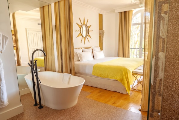 A hotel room with a bathtub, a bed with a yellow blanket, a sun-shaped mirror, and a window with curtains.