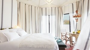 A bedroom with a large bed, a wooden chair, a small table with a fruit bowl, and a view of the outdoors through curtains. - Brindos Lac & Château (Anglet)