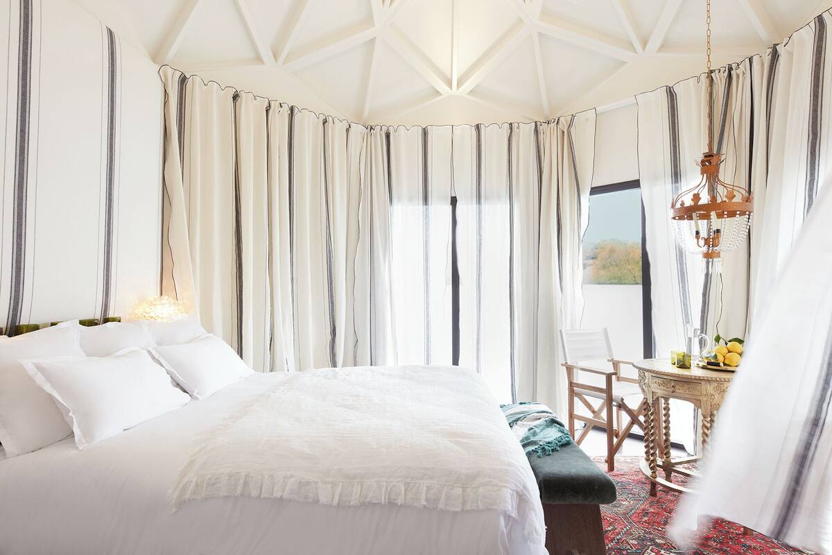 A bedroom with a large bed, a wooden chair, a small table with a fruit bowl, and a view of the outdoors through curtains.