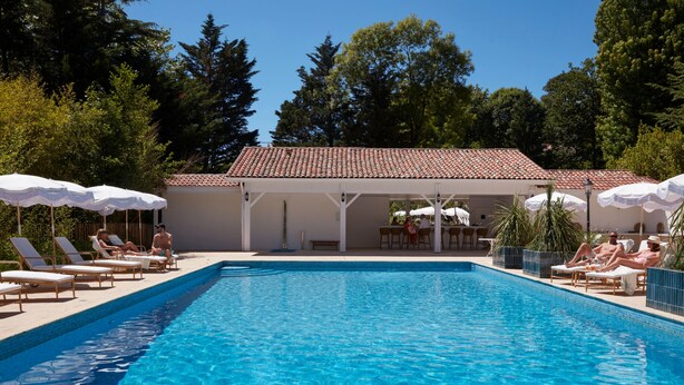 Una piscina con sillas de descanso y sombrillas, rodeada de árboles y un edificio con techo de tejas.