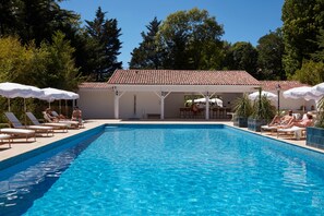 A swimming pool with lounge chairs and umbrellas, surrounded by trees and a building with a tiled roof.