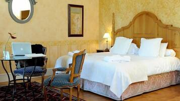 A hotel room with a bed, a desk with a laptop, two chairs, and a bedside table with a lamp.