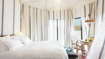A bedroom with a large bed, a wooden chair, a small table with a fruit bowl, and a view of the outdoors through curtains.