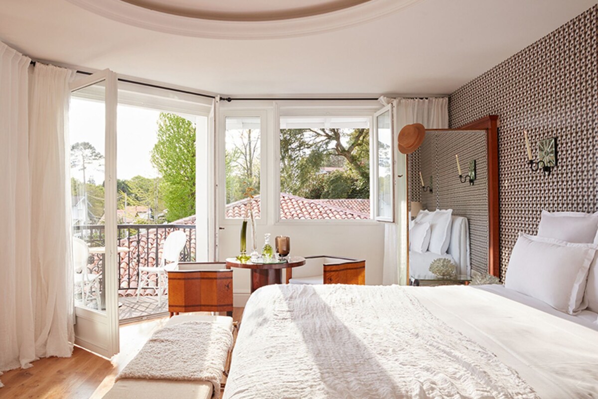 A bedroom with a large bed, a small table, and a balcony with a view of the outside.