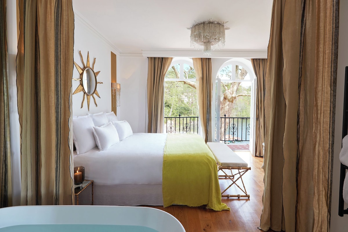 Habitación de hotel con cama, mesita de noche con una vela, ventana con cortinas y vistas al exterior.
