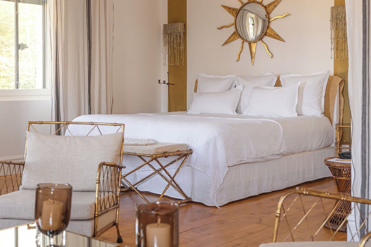 A bedroom with a large bed, a sun-shaped mirror, a window with curtains, and a patterned rug.