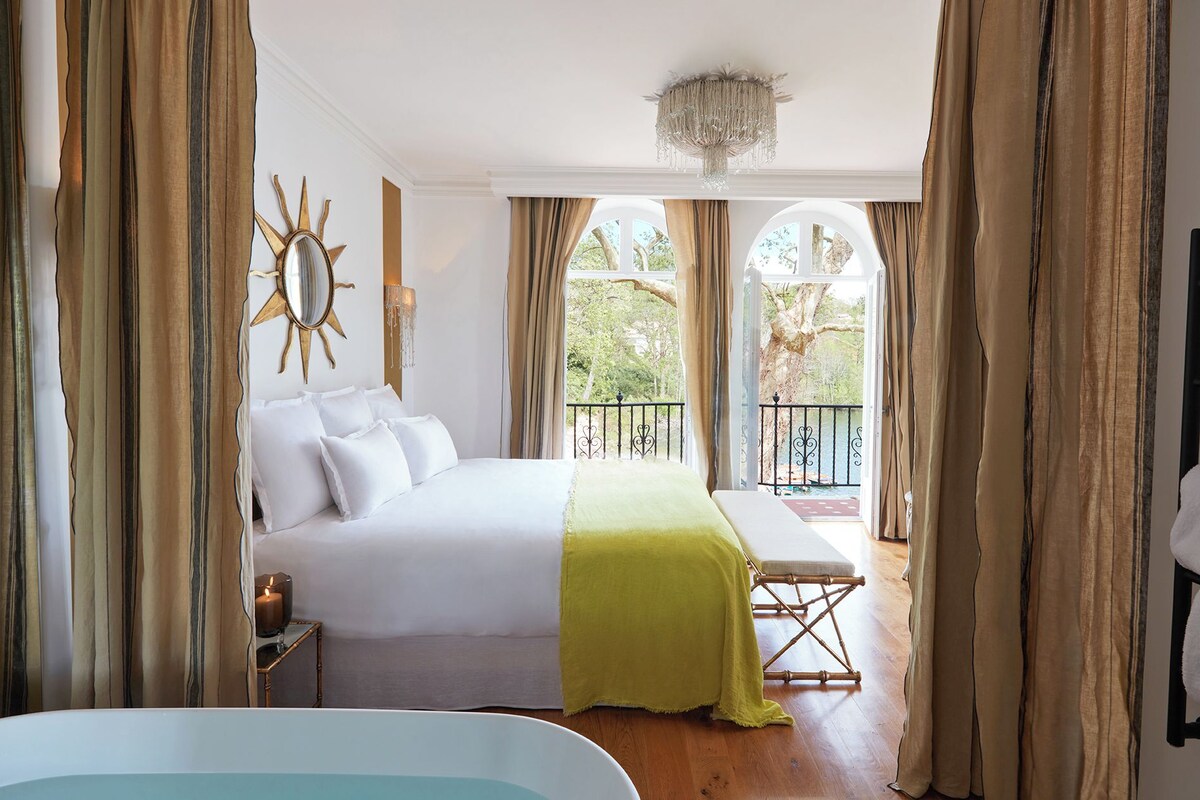 A hotel room with a bed, a bedside table with a candle, a window with curtains, and a view of the outdoors.