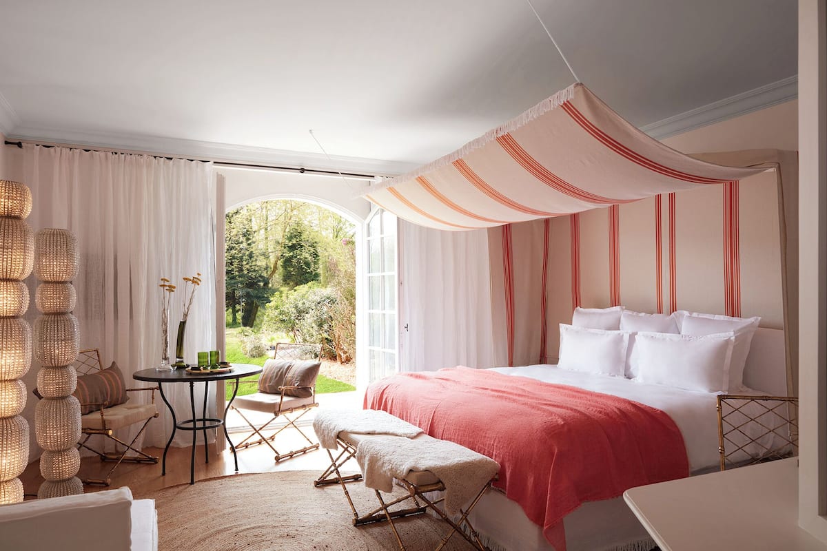 A bedroom with a large bed, a small table, and a chair. There is a window with curtains, a wicker lamp, and a view of greenery outside.