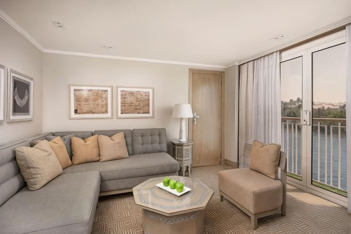 A living room with a sofa, a coffee table with fruit, a lamp, and a view of the outdoors through large windows.