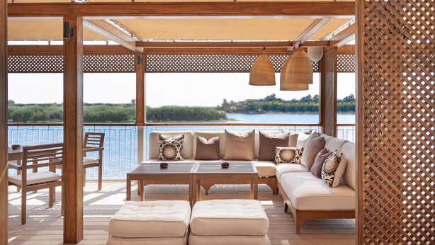 A wooden deck with a sofa, ottomans, and a table, overlooking a body of water. - JAZ  Elite Viceroy Nile Cruise - Every Saturday from Luxor for 07 & 04 Nights - Every Wednesday From Aswan for 03 Nights (Luxor)