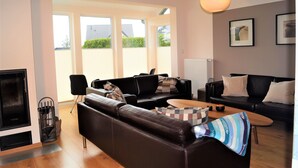 A modern living room with a fireplace, two black sofas, a wooden coffee table, and a large window offering a view of the outside.