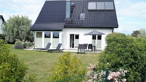 A house with a sloped roof, a patio with a table and chairs, and a lawn with bushes and flowers.