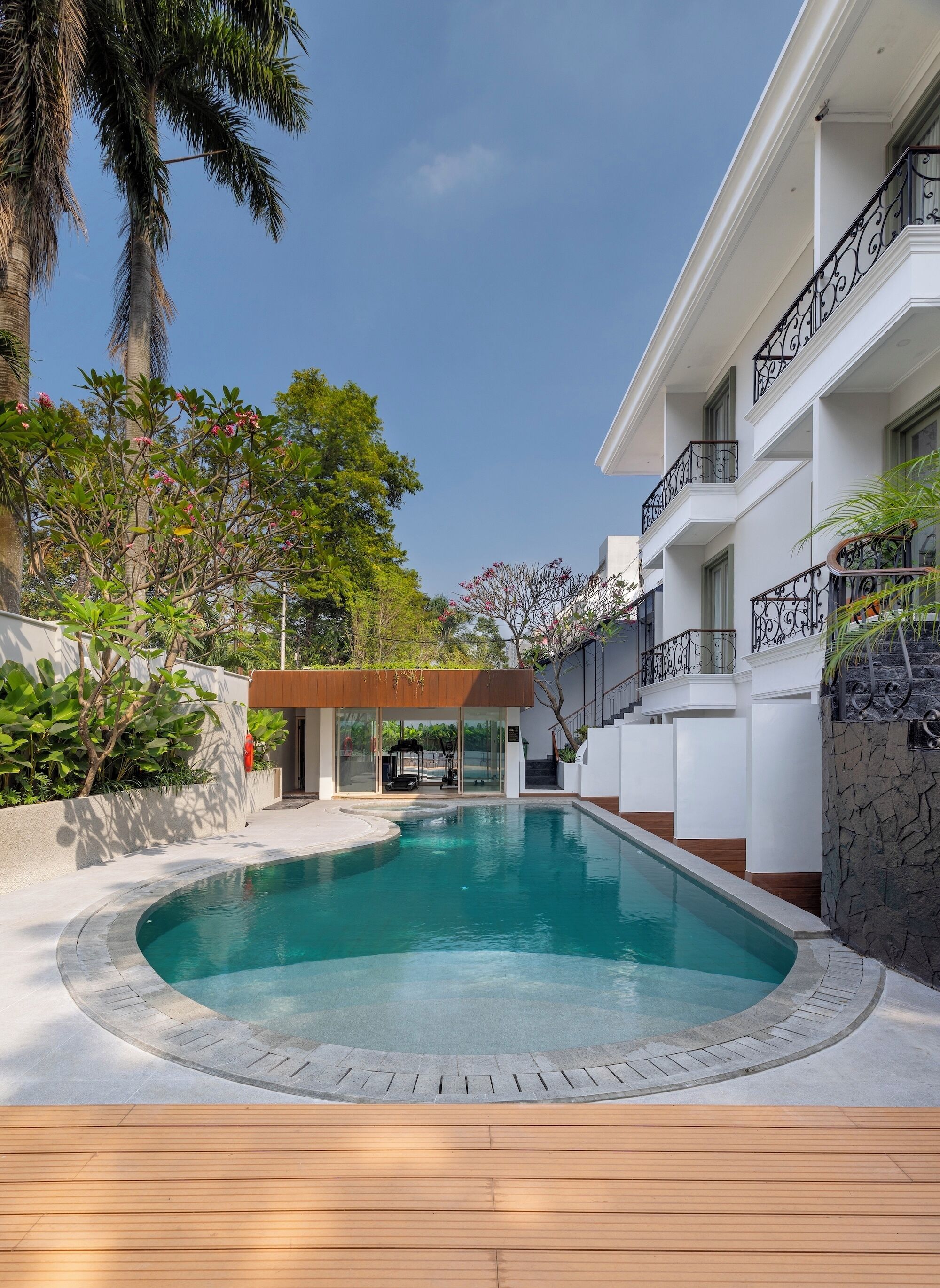 A swimming pool with a wooden deck, surrounded by a white building with balconies and a stone wall.