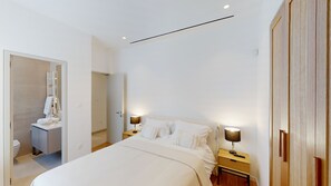Una camera da letto moderna con un letto grande, comodini con lampade, un'armadio in legno e un bagno visibile attraverso una porta aperta.