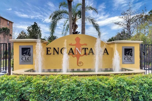 A yellow sign with 'ENGANTA' and a lizard logo, flanked by two fountains, with palm trees and a clear sky in the background.