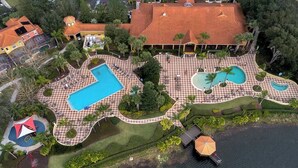An aerial view of a resort with multiple swimming pools, a large building with a red-tiled roof, and a body of water. - Encantada - 4 bed 3 bath lakefront corner house (Kissimmee)