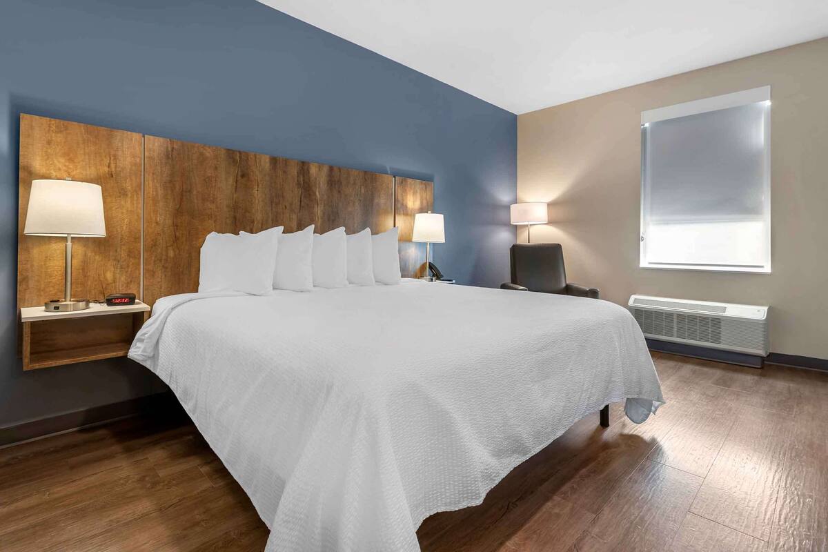 A hotel room with a large bed, two bedside tables with lamps, a wooden headboard, a window with blinds, and a wall-mounted heater.