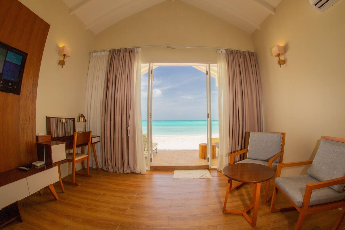 A room with a wooden floor, a desk with a lamp, two chairs, and a view of the beach through a doorway.
