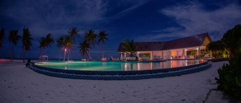 A beachfront hotel with a swimming pool, palm trees, and a thatched-roof building.