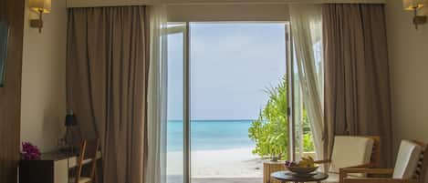 A room with a desk, chairs, a small table with a bowl of fruit, and a view of the beach through a glass door.