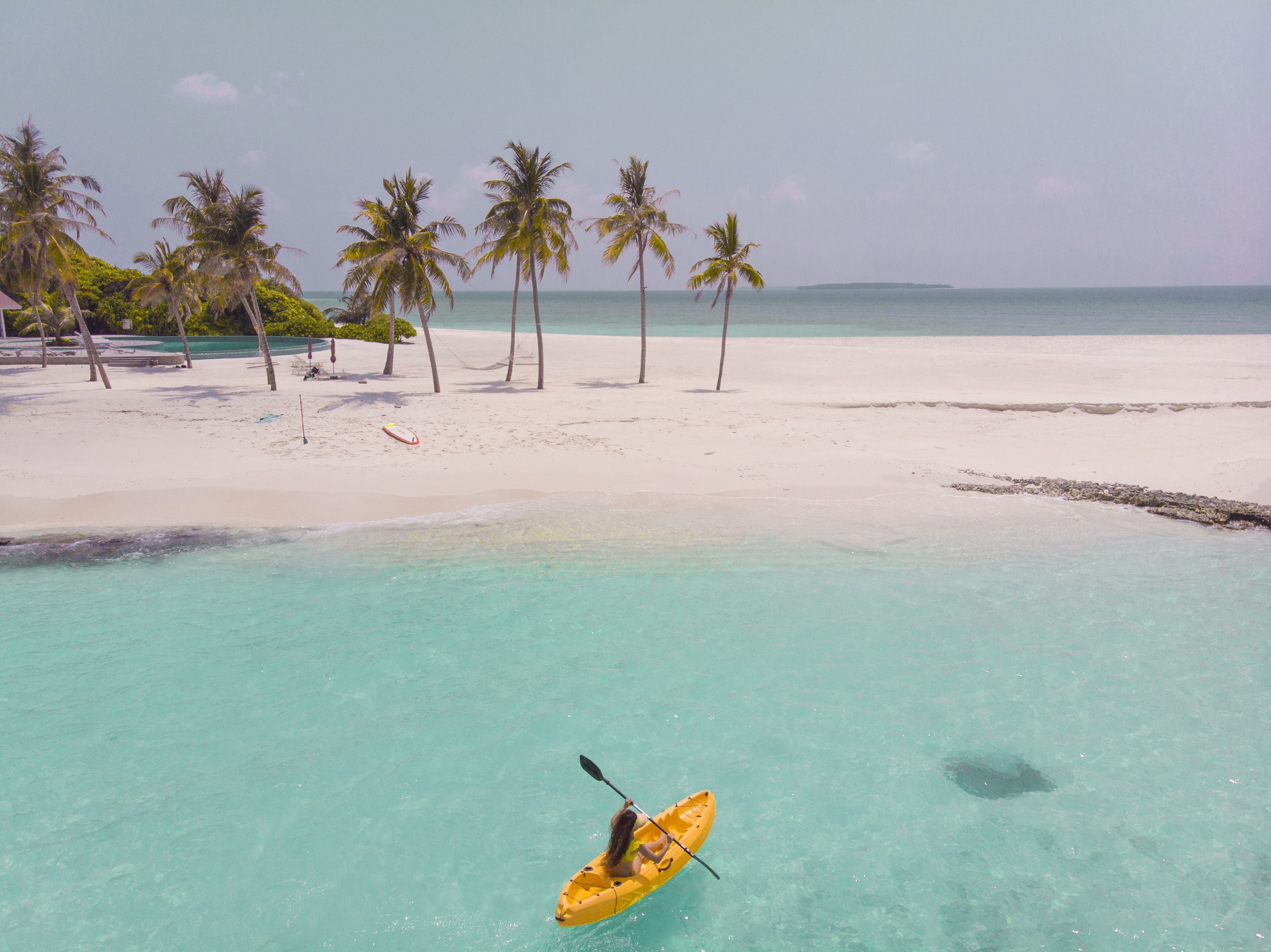A beach with clear turquoise water, a person kayaking, and palm trees.