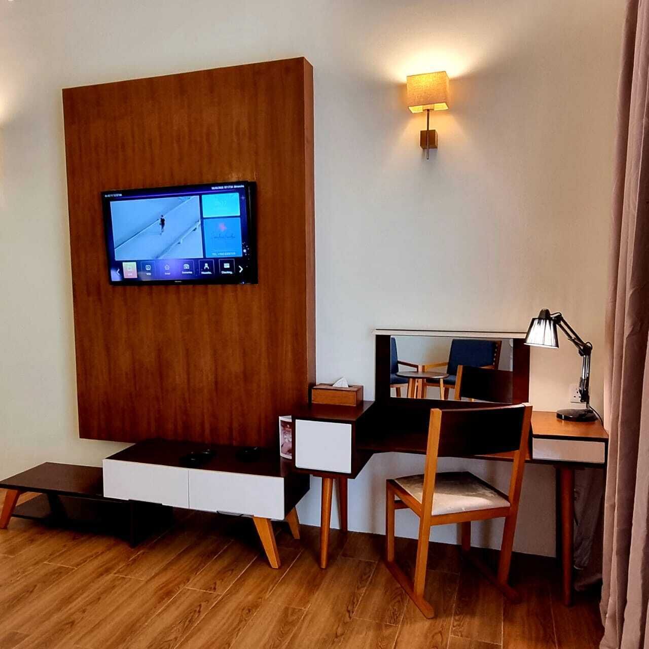 A hotel room with a wooden panel wall, a mounted television, a desk with a chair, a lamp, and a bedside table.