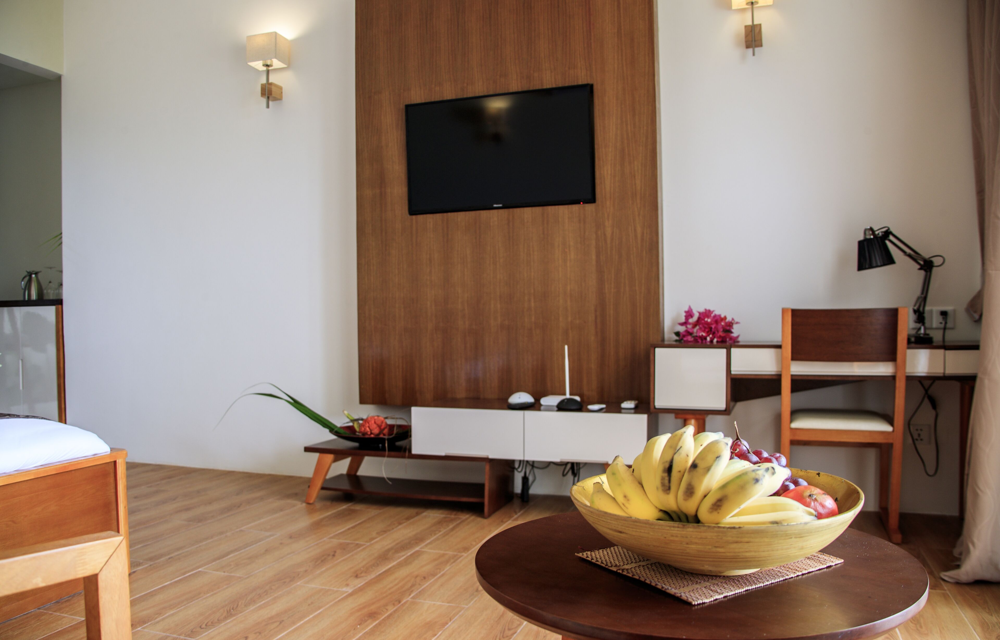 Una habitación de hotel moderna con suelo de madera, un televisor de pantalla plana en la pared, una mesa de comedor de madera con un cuenco de fruta y un escritorio con una lámpara.