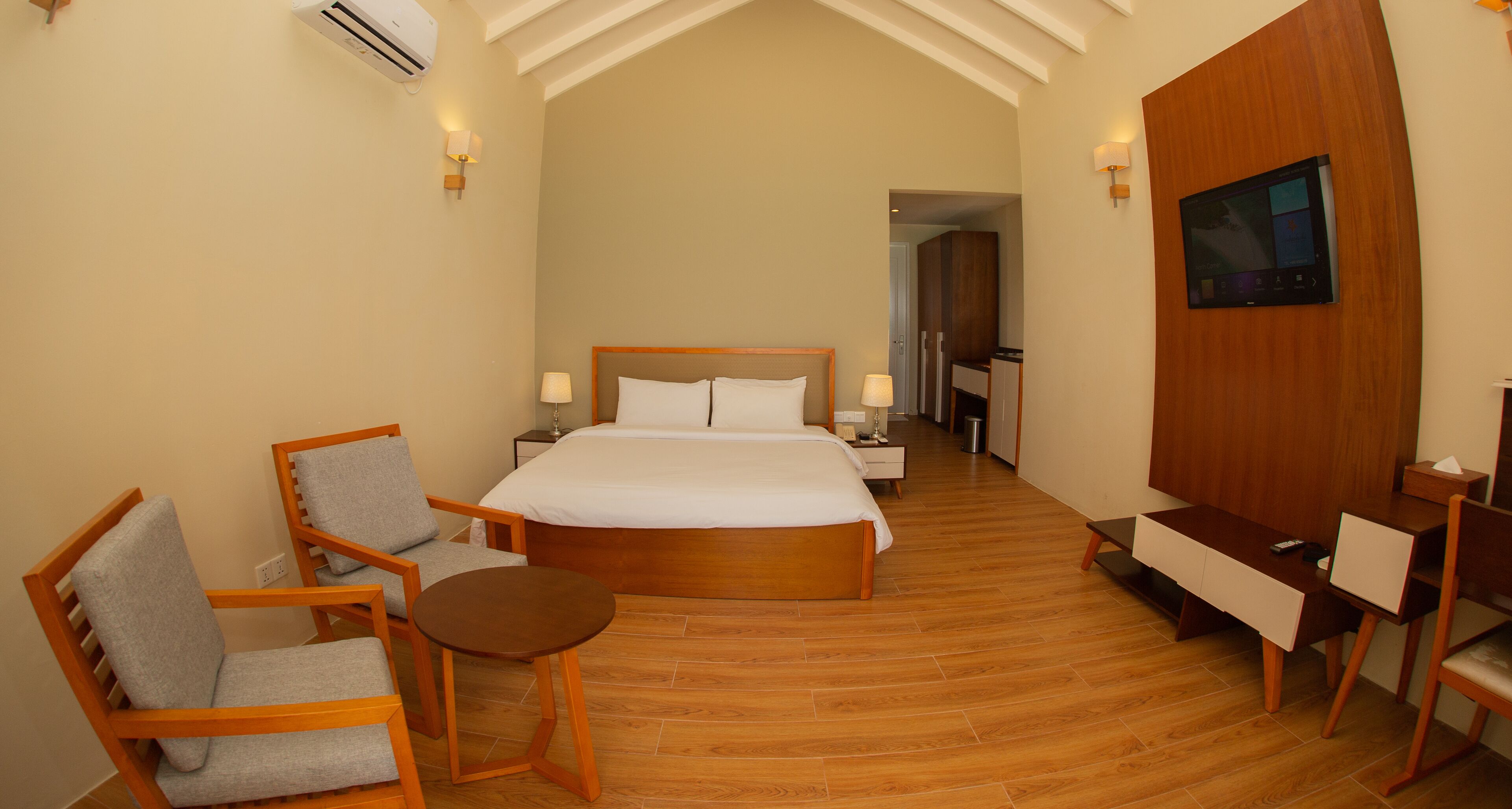 A hotel room with a large bed, a wooden desk, a chair, a small table, and a TV.