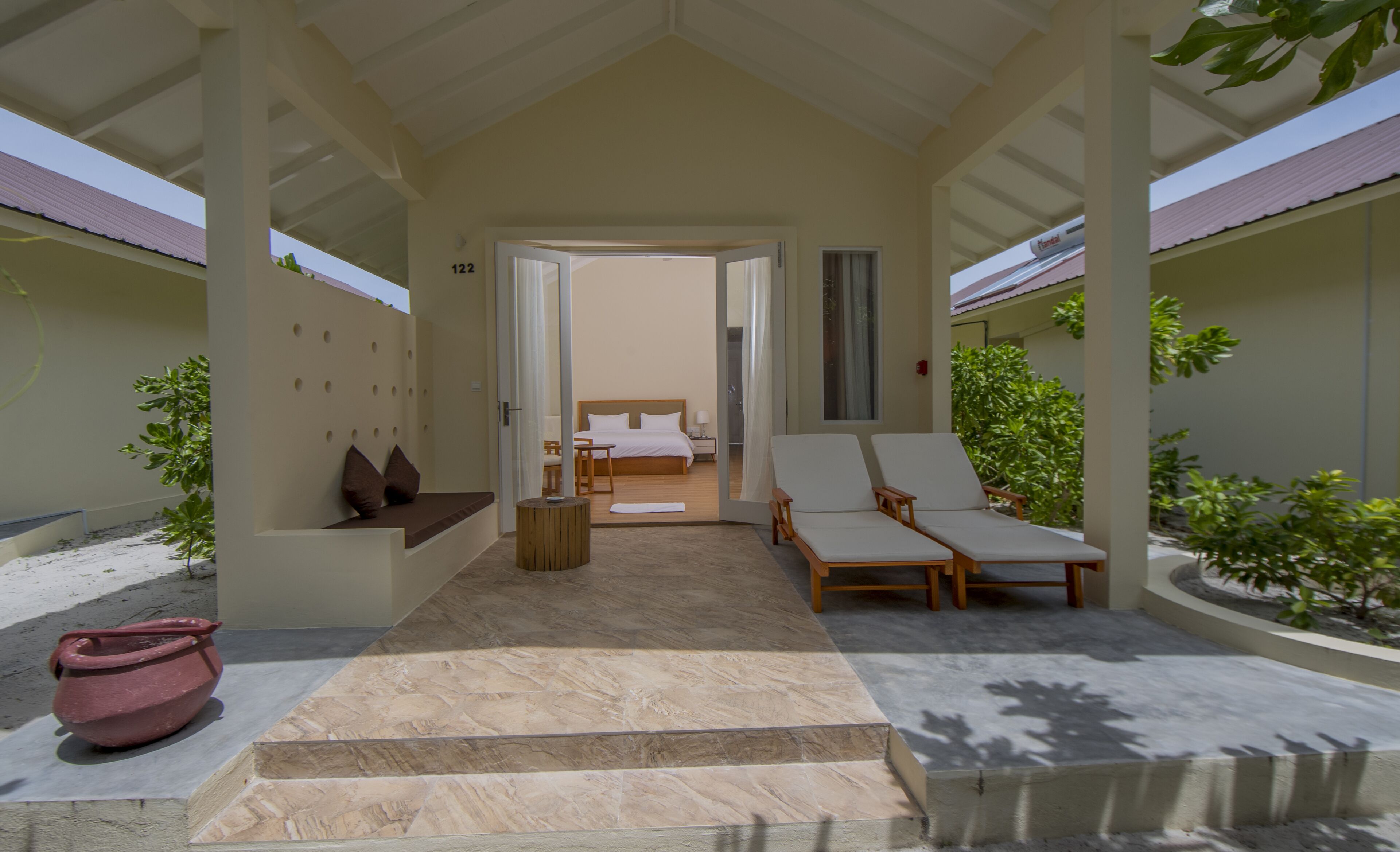 A covered outdoor seating area with a bed and lounge chairs, a potted plant, and a tiled floor.