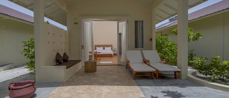 A covered outdoor seating area with a bed and lounge chairs, a potted plant, and a tiled floor.