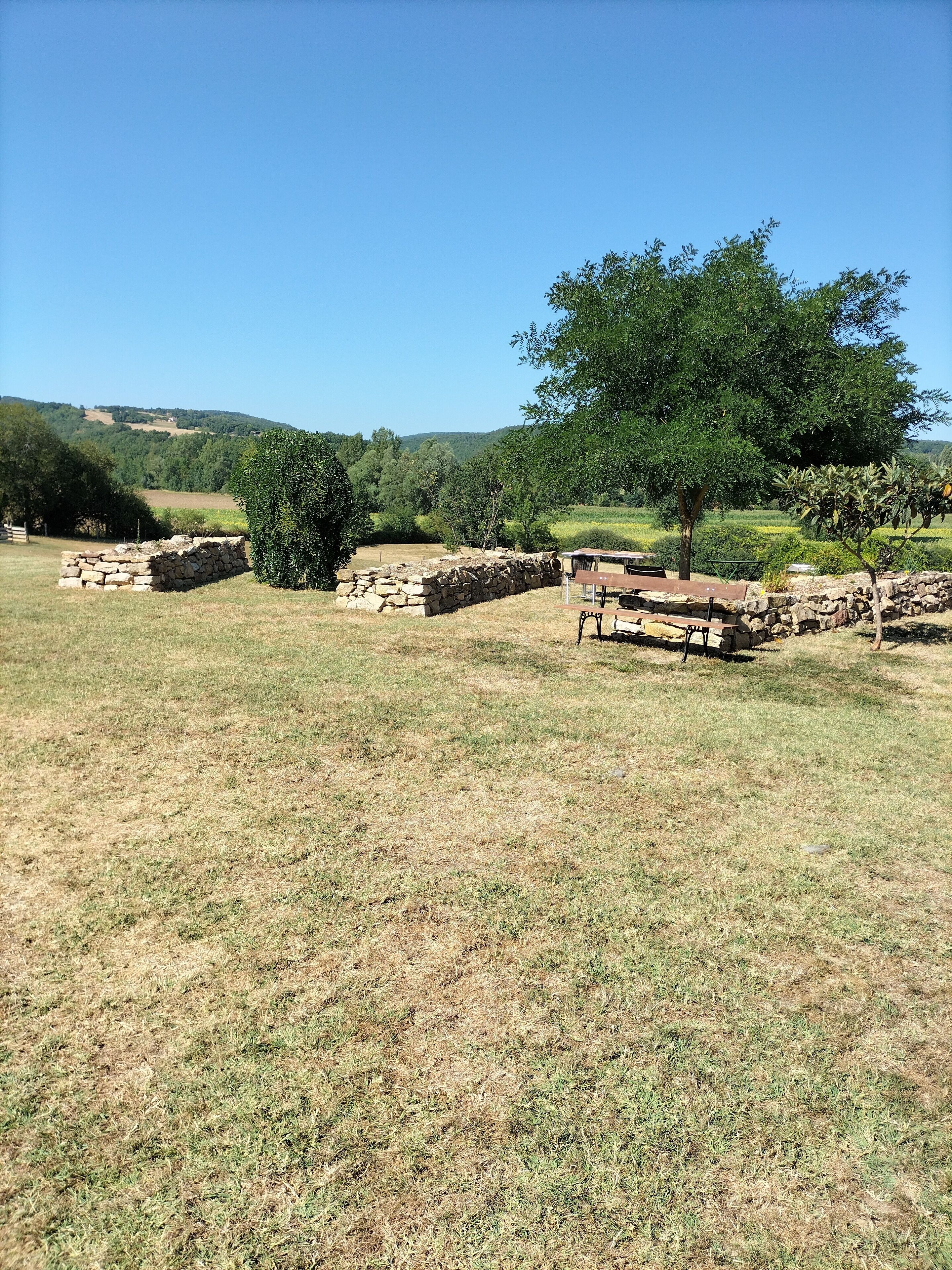 A stone structure with a bench and a table in a grassy field with trees and hills in the background.