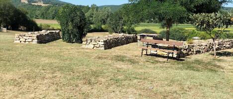 A stone structure with a bench and a table in a grassy field with trees and hills in the background.