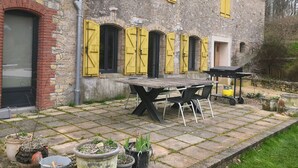 Een rustiek stenen gebouw met gele luiken, een bakstenen ingang en een verhard terras met tuinmeubilair.