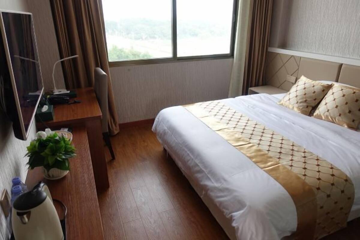 Quarto de hotel com cama, escrivaninha, cadeira, planta em vaso e janela com cortinas.