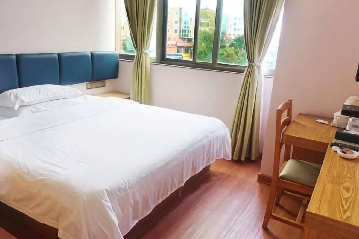 Quarto de hotel com cama, escrivaninha com cadeira e janela com cortinas.