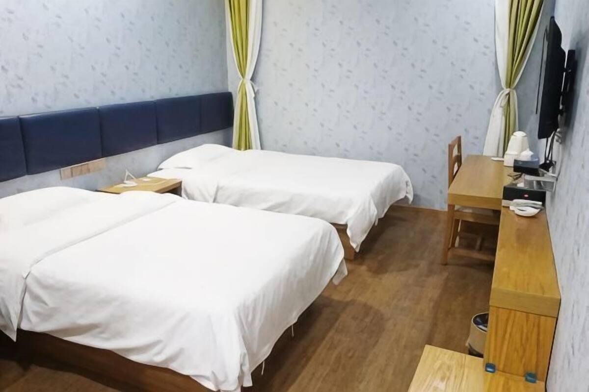 Quarto de hotel com duas camas, uma escrivaninha de madeira, uma cadeira, uma televisão e uma mesinha com abajur.