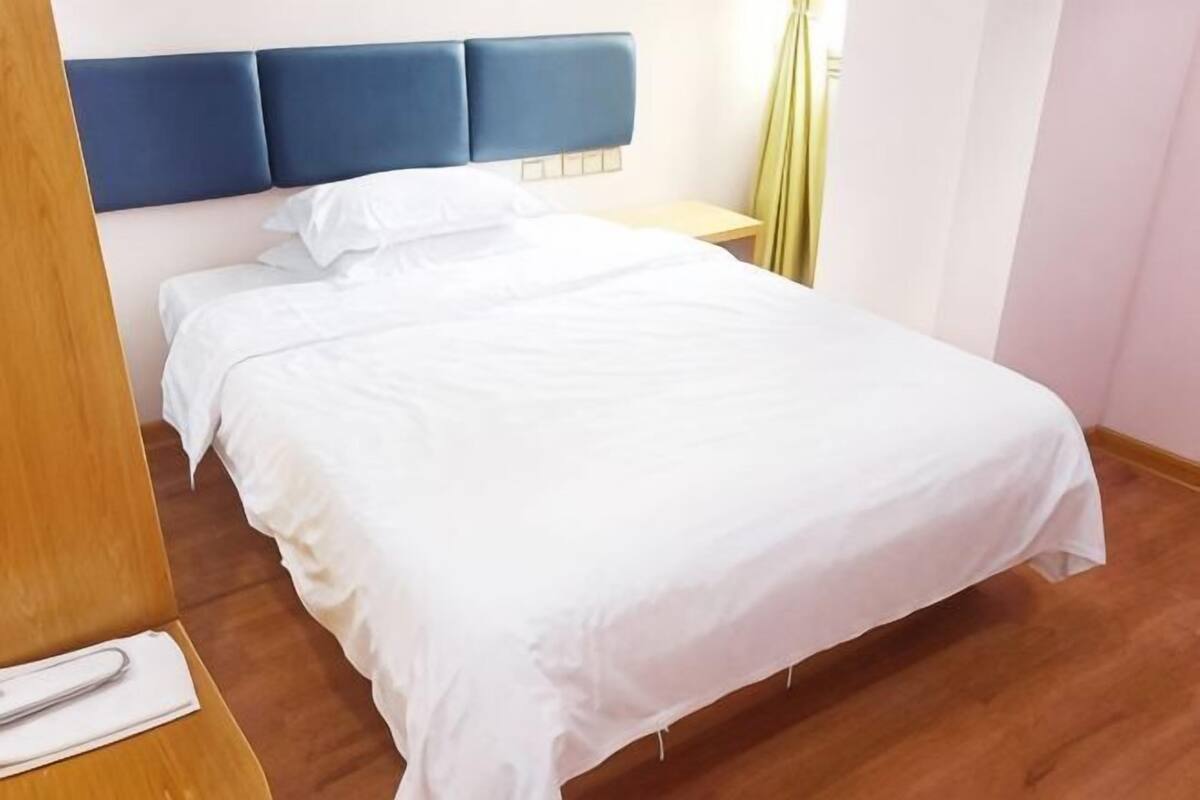 Uma cama com roupa de cama branca, cabeceira azul, mesa de cabeceira de madeira e cortina amarela.