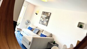 Een moderne woonkamer met een bank, een ventilator, een kleine tafel met een plant en een aan de muur hangend schilderij.