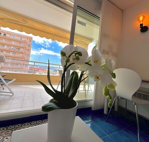 Uma varanda com uma orquídea em vaso, uma cadeira branca e vista para um edifício com varandas.