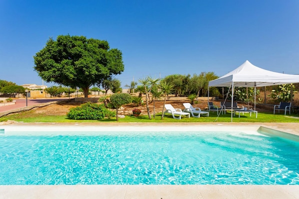 Una piscina con agua azul cristalina, rodeada de sillones blancos y un toldo también blanco.